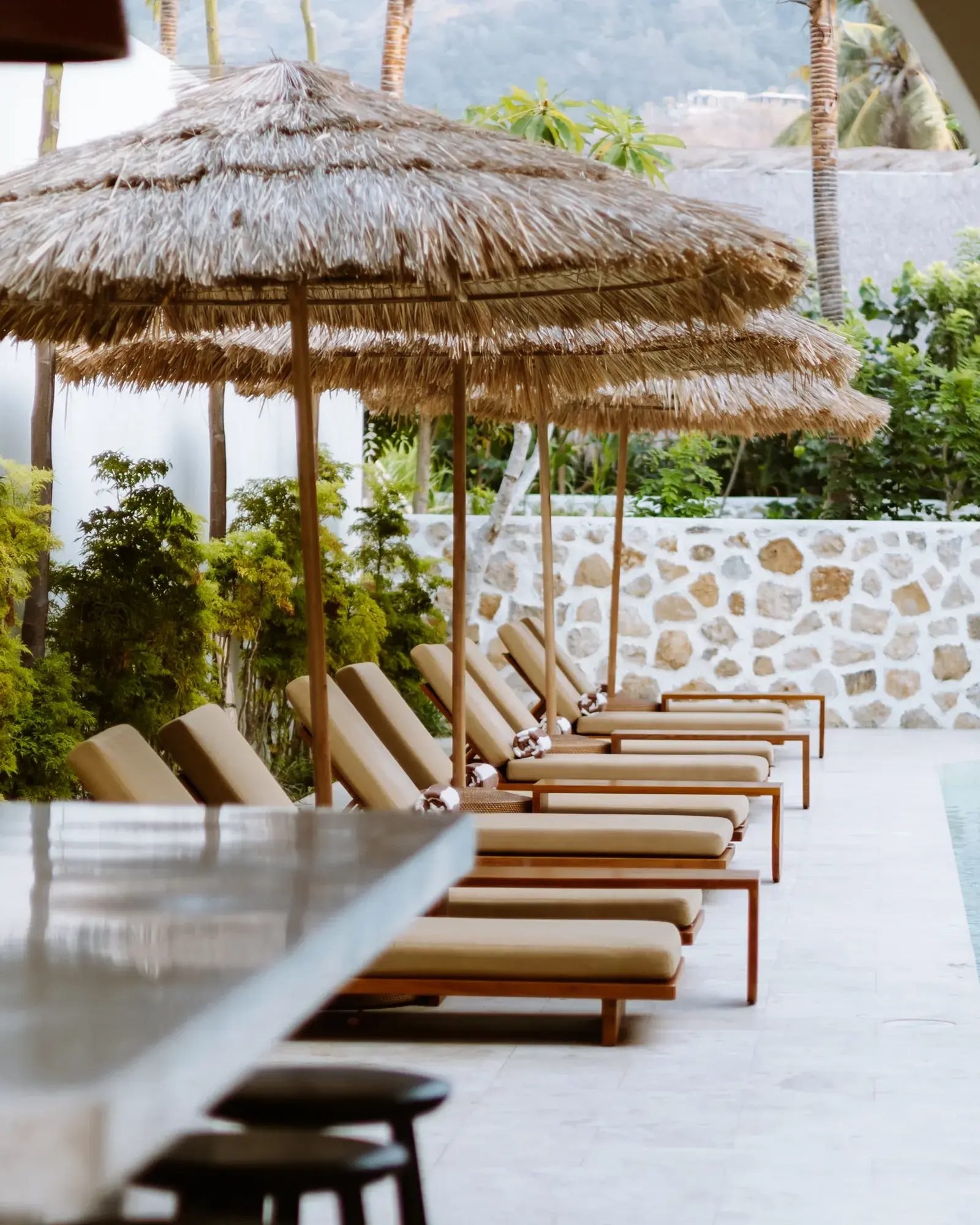 Cushioned sun loungers beneath thatched parasols beside the Boni Beach hotel pool, with tropical plants and a stone wall.