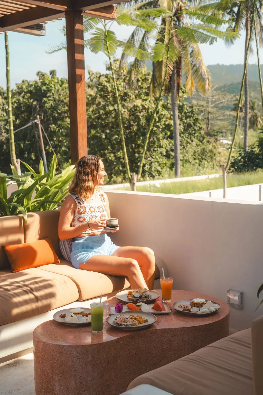 A guest with coffee beside breakfast dishes and fresh juices in Boni Beach’s upstairs rooftop dining area, overlooking palm trees and hills.