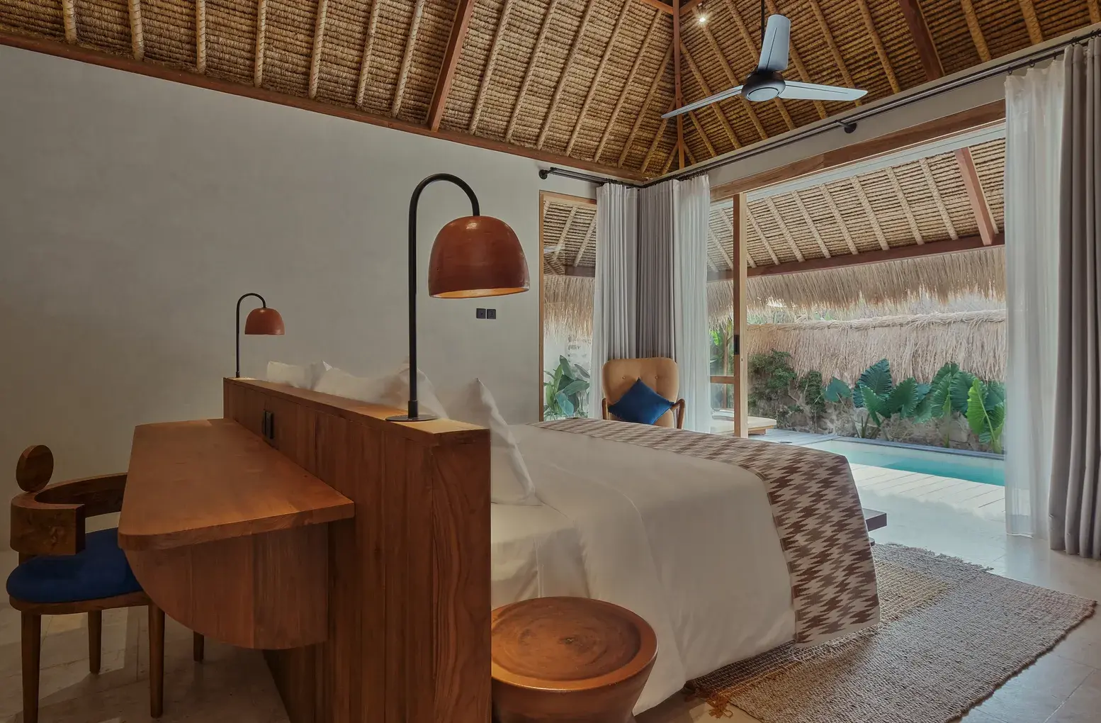 A Deluxe Pool Bungalow bedroom at Boni Beach Lombok, with teak furniture, a thatched ceiling and its private pool visible through glass doors.