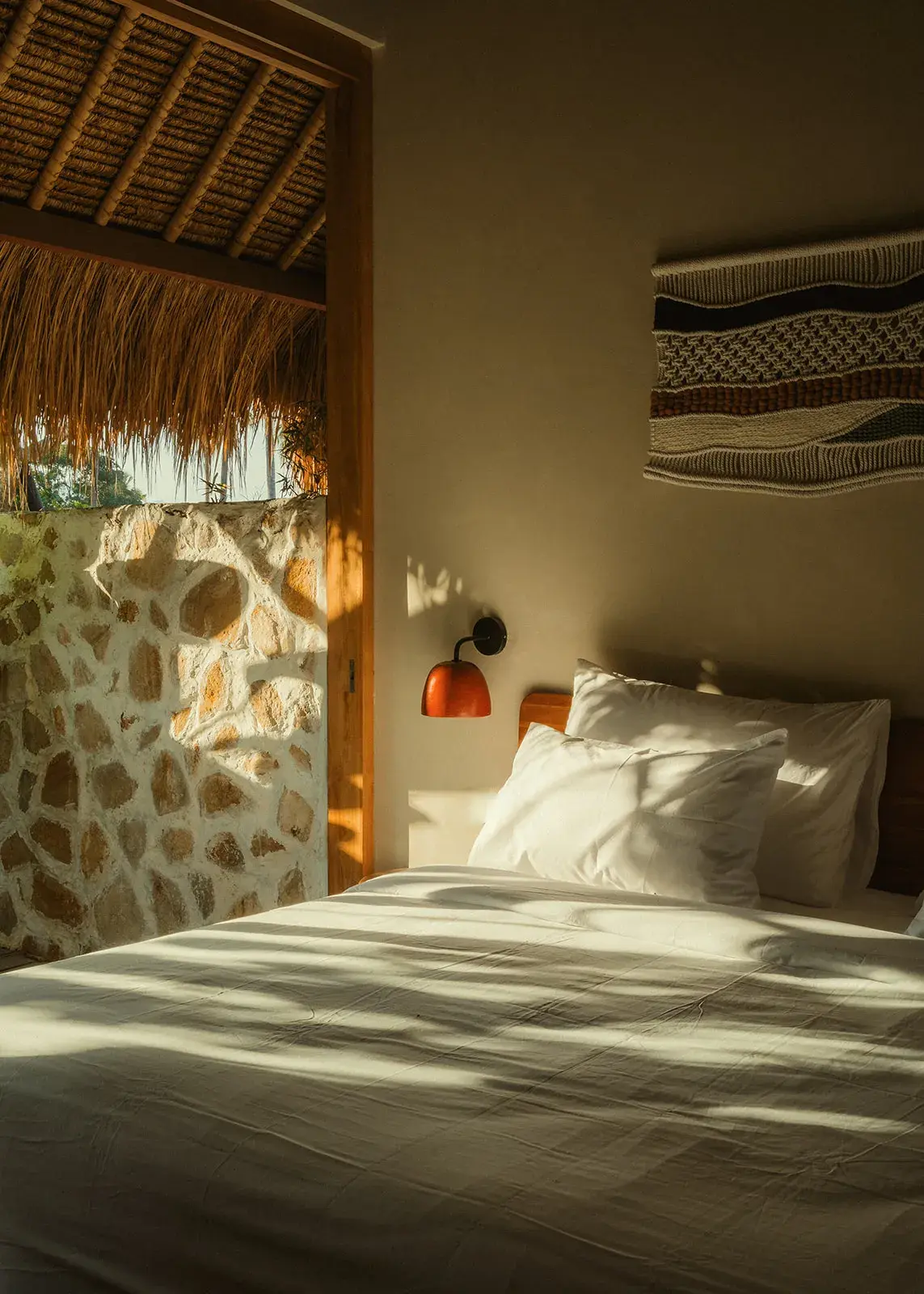 Afternoon sunlight across white bedding and a teak headboard, with macramé artwork on the villa bedroom wall.
