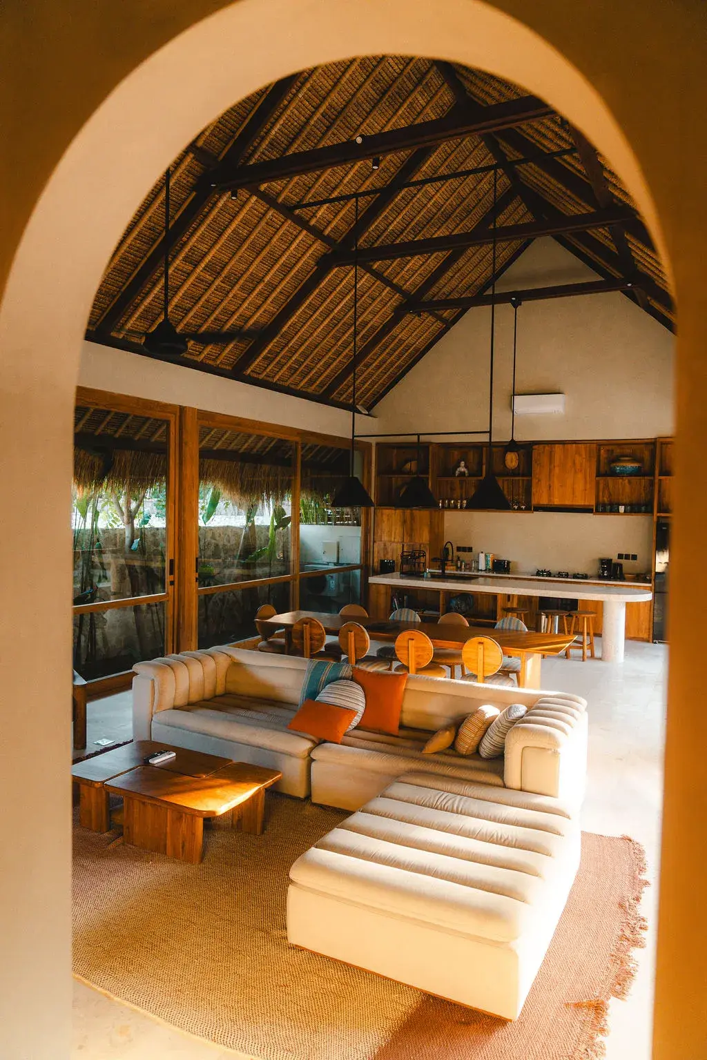 Villa lounge viewed through an archway, with a cream sofa, teak dining table and kitchen beneath a thatched ceiling.