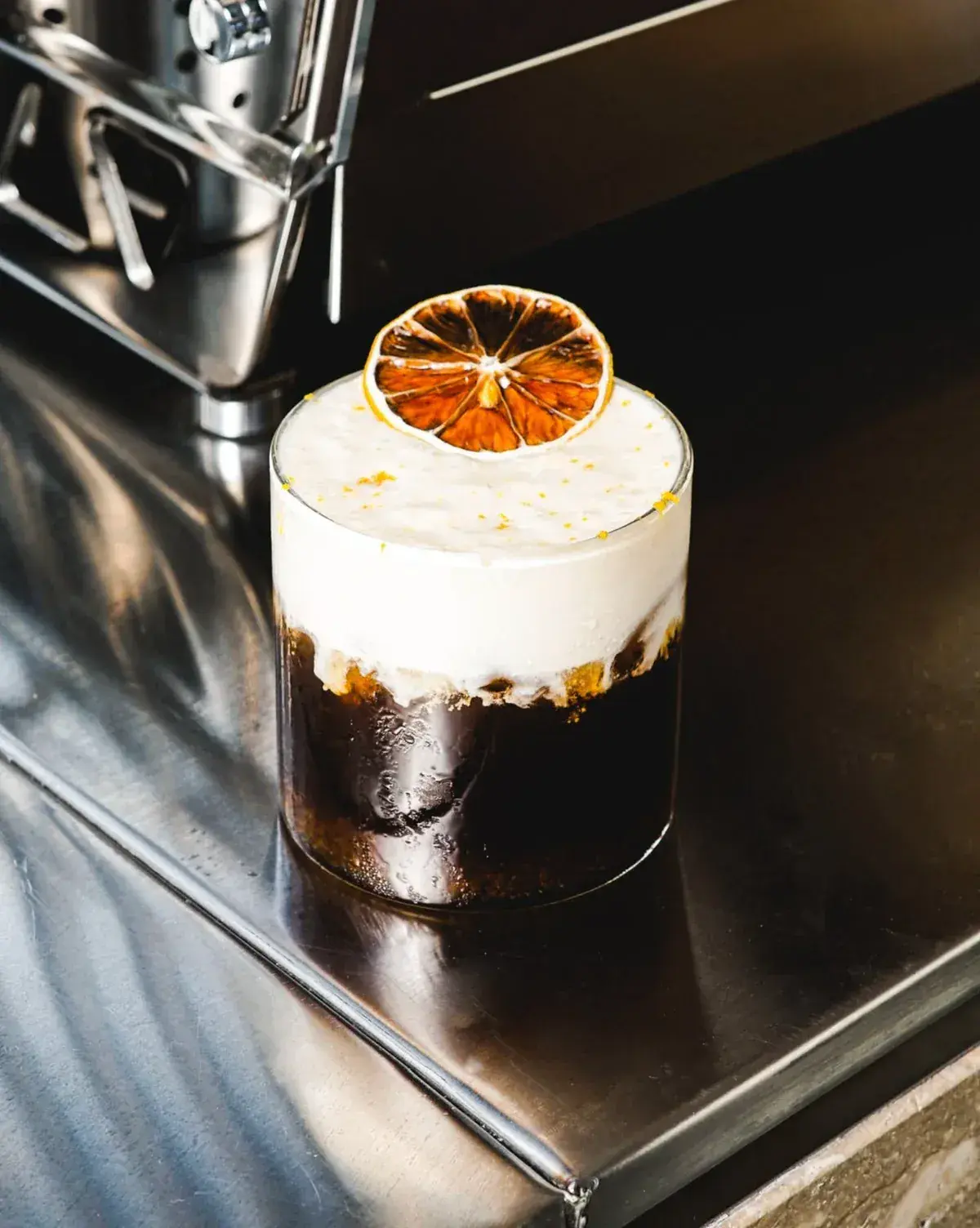 Sookla’s Mont Blanc coffee with a thick cream layer and dried orange, beside an espresso machine.